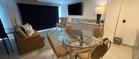 Una sala de estar moderna con una mesa de comedor de vidrio, sillas acolchadas, un sofá marrón y un ventanal con cortinas oscuras.