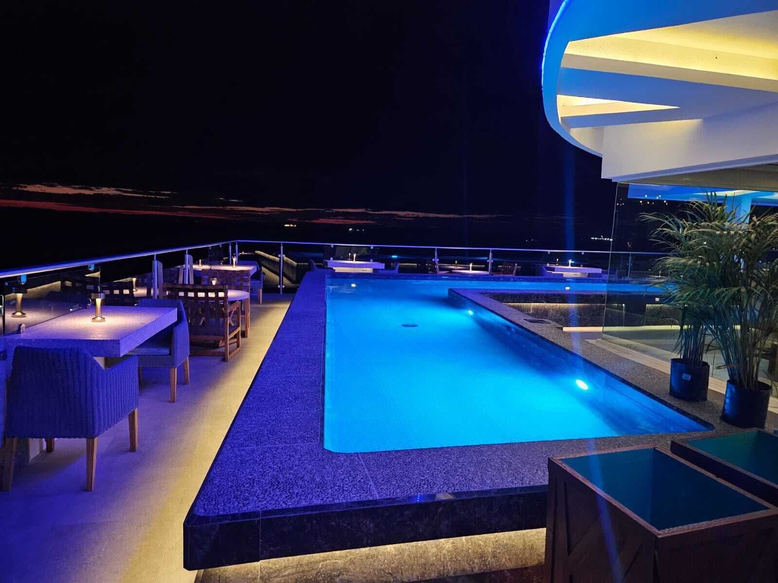 Area piscina sul tetto con tavoli e sedie, una piscina e vista sulla città di notte.