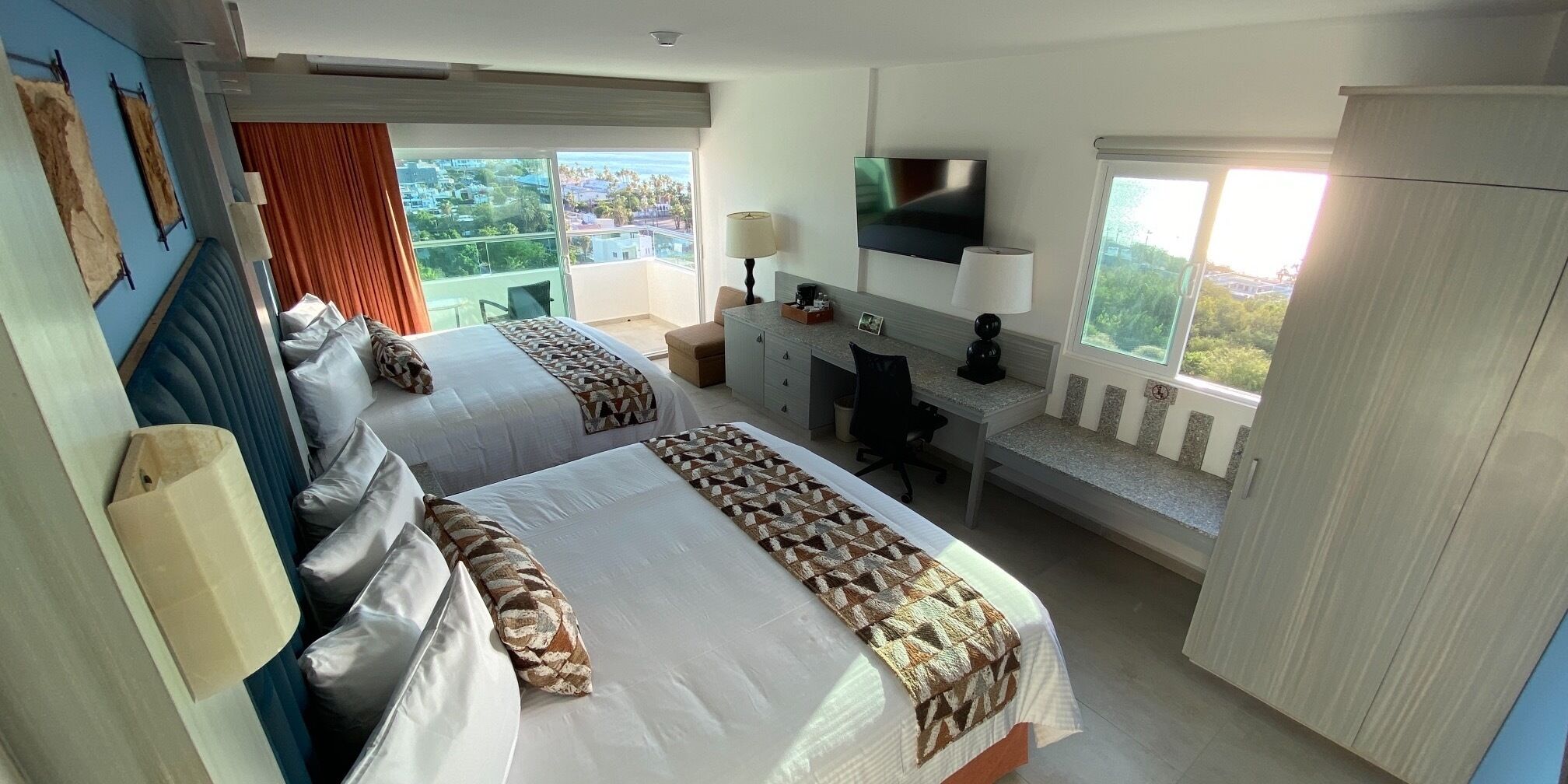 A hotel room with two beds, a desk, a TV, and a balcony with a city view.