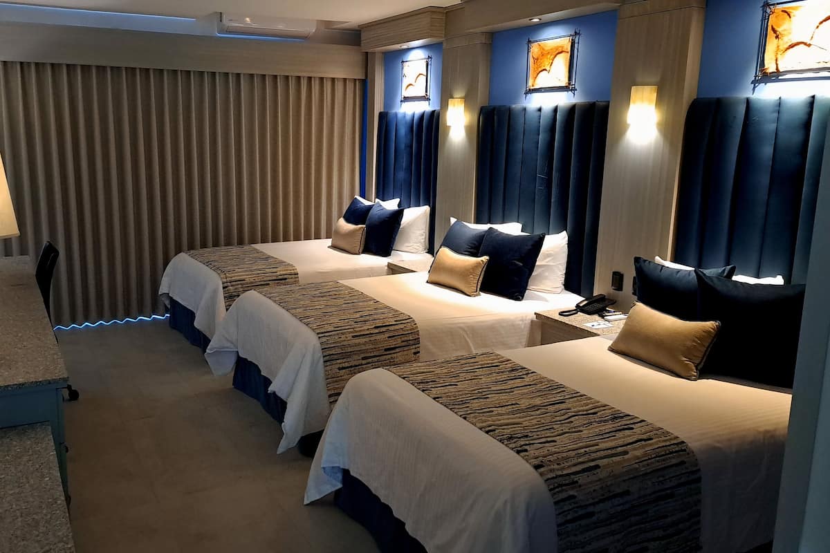 Une chambre d’hôtel avec trois lits, un bureau, une lampe et une fenêtre avec des rideaux.