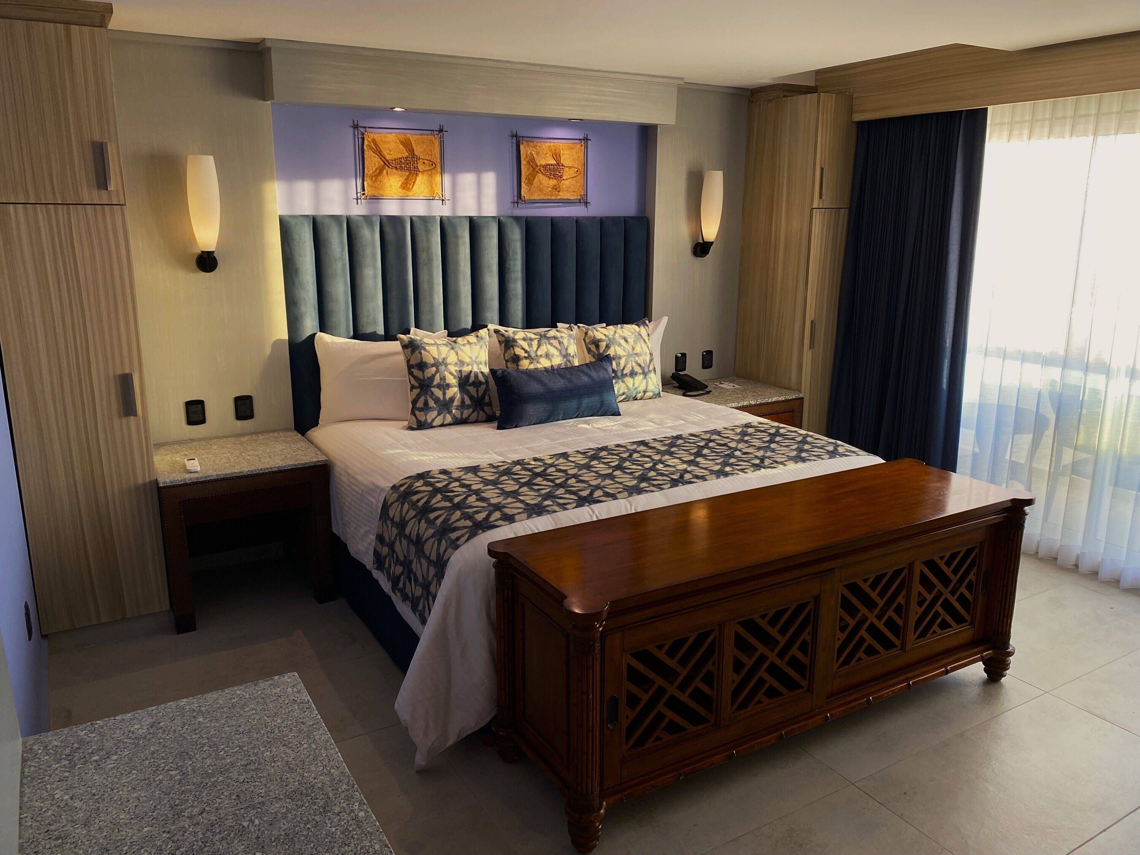 A modern hotel room with a large bed, a wooden headboard, two wall-mounted lamps, and a view of the outdoors through sheer curtains.