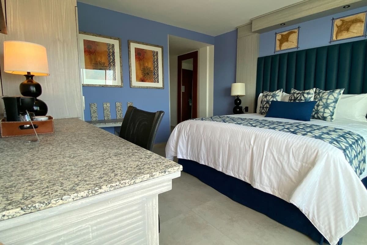 Une chambre d’hôtel avec un lit, un bureau et une chaise. Il y a deux lampes sur le bureau et une porte qui donne sur une autre pièce.