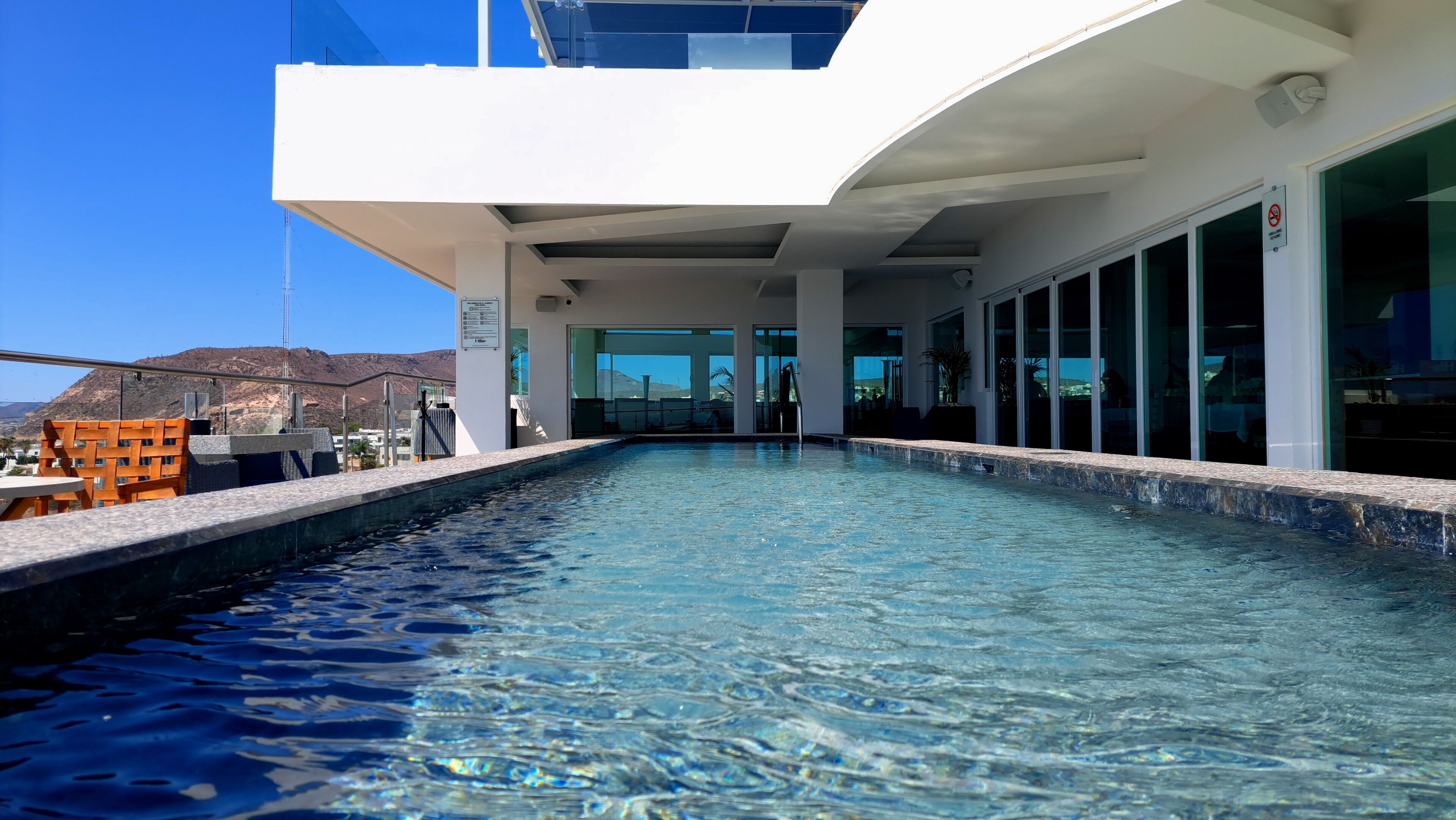 Uma área de piscina moderna com água azul cristalina, um edifício branco com grandes janelas e vista para a paisagem circundante.