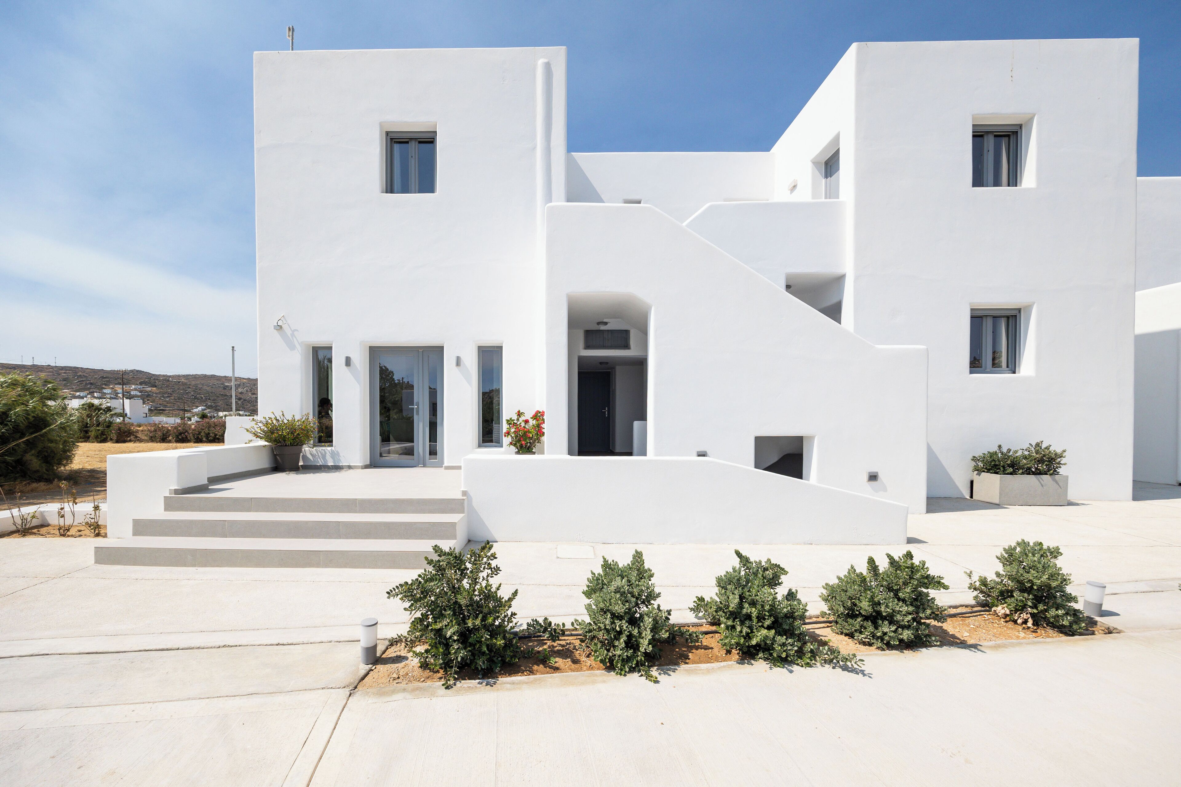 Un edificio moderno blanco con techo plano, entrada central y un pequeño jardín con arbustos.