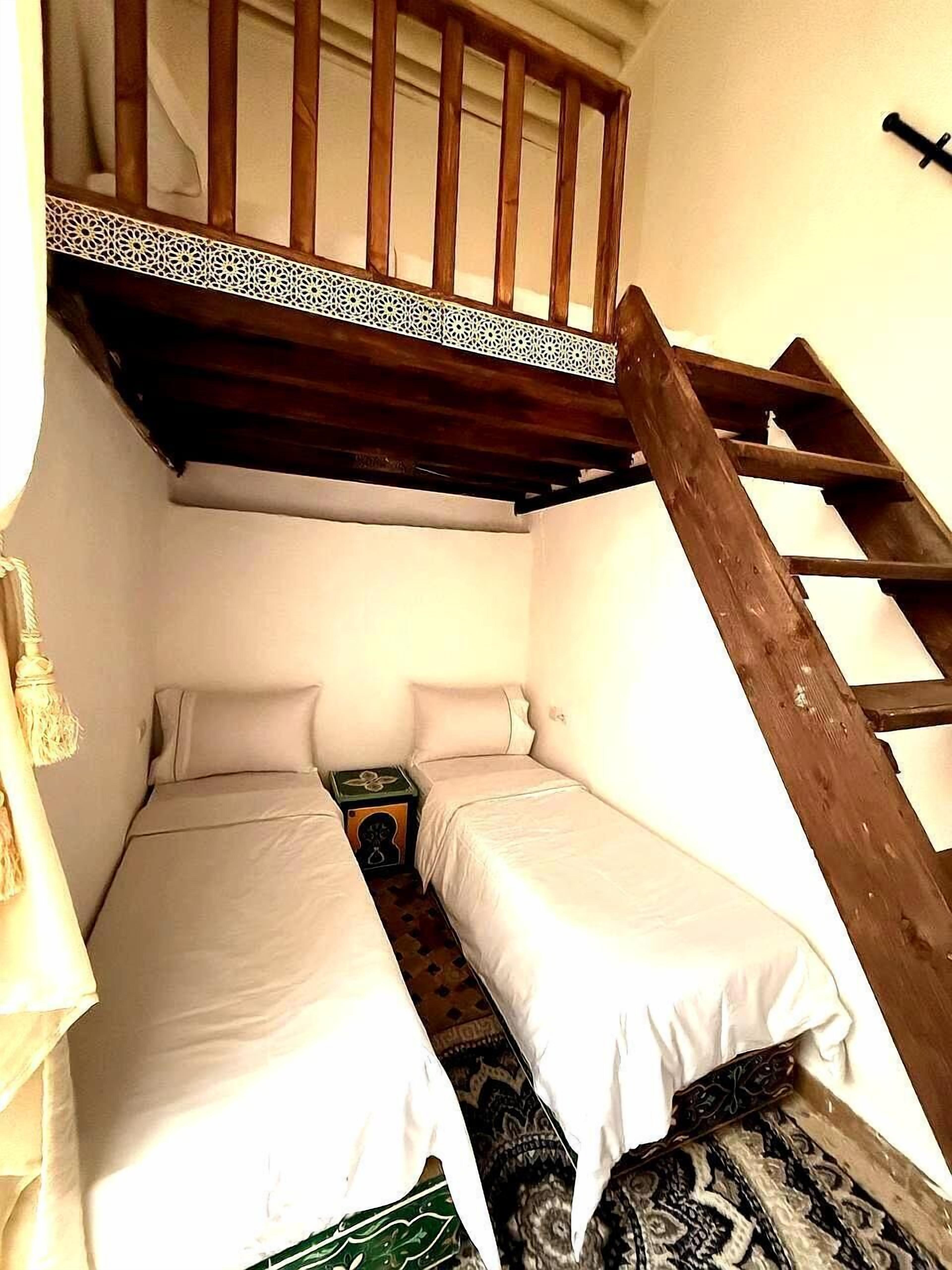 A small, compact room with two single beds, a wooden ladder, and a small table with a decorative item.