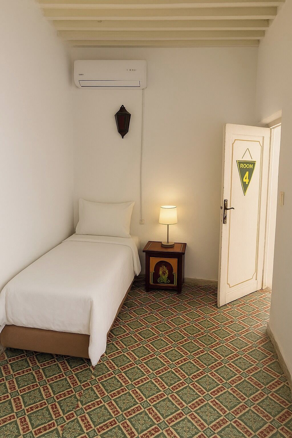 Une petite pièce, bien rangée, avec un lit, une table de chevet dotée d’une lampe, une porte portant l’inscription « CHAMBRE 4 » et un sol à motifs.