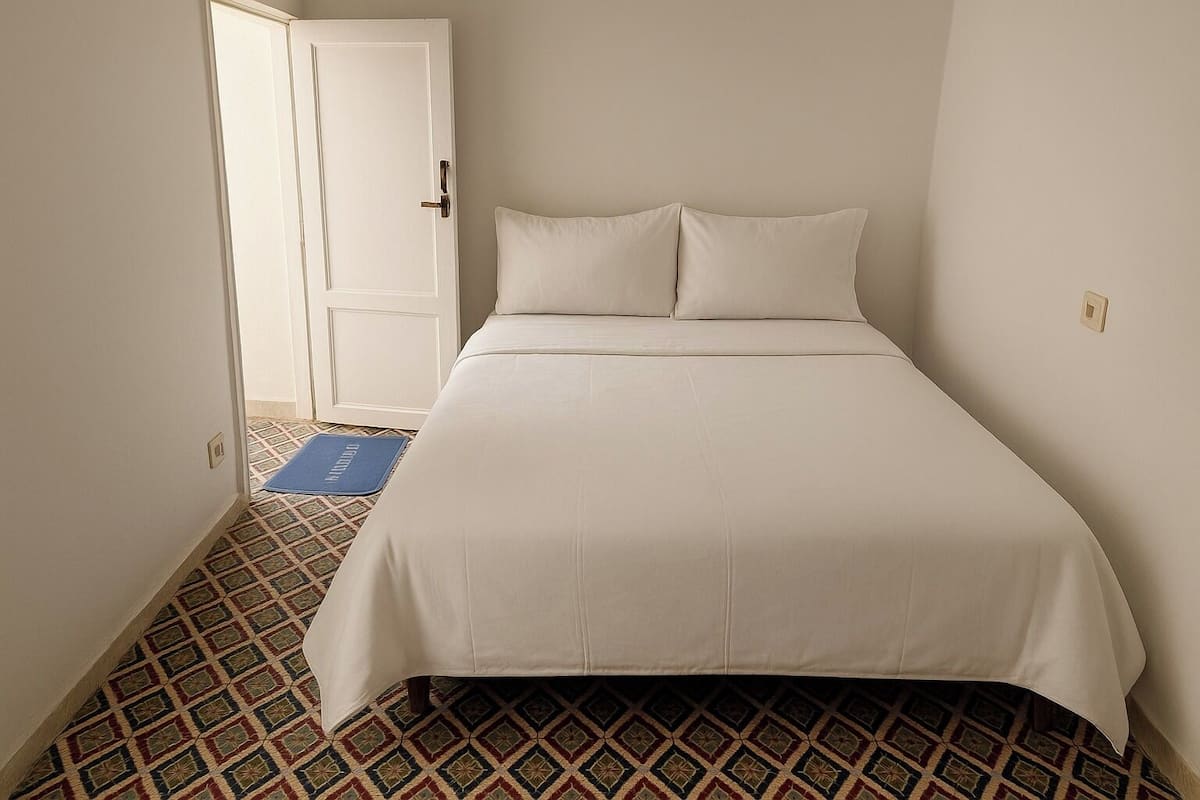 Habitación con una cama individual, suelo estampado, colcha blanca y felpudo azul en la entrada.