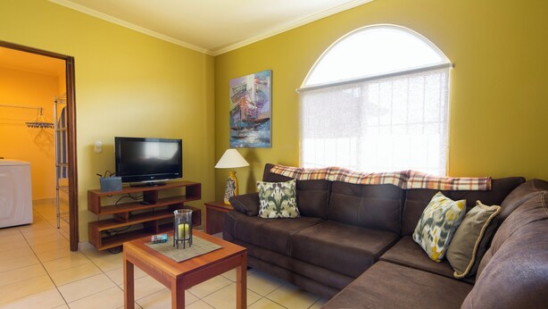 A living room with a brown leather sofa, a television on a wooden stand, a lamp, and a painting on the wall. - 2-bedroom apartment in charming Cuidad Colon with WiFi. (Cuidad Colon)