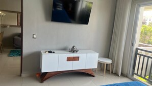 Ein modernes Hotelzimmer mit Flachbildfernseher an der Wand, einem weißen Schminktisch mit Schubladen, einem kleinen runden Tisch und einem großes Bett mit blauer Bettdecke.