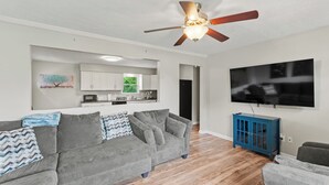 Ein modernes Wohnzimmer mit grauem Sofa, Deckenventilator, Flachbildfernseher und einer Küche im Hintergrund.