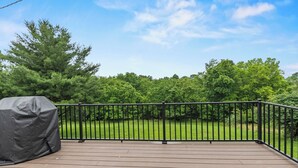 Eine Holzterrasse mit schwarzer Metallreling, einem Grill und Blick auf eine grüne Rasenfläche und Bäume.