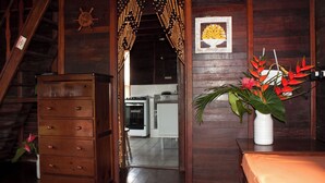 Un intérieur en bois avec un escalier, un meuble blanc et une cuisine visible à travers une porte ouverte.
