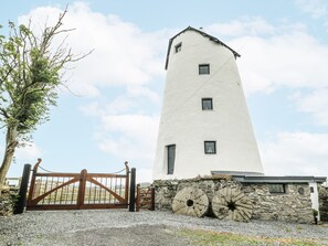 A tall, white, cylindrical building with a stone base and a wooden gate in front. - The Old Cornmill (Cemaes Bay)