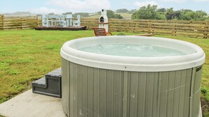 A hot tub in a grassy outdoor area with a wooden deck, a white gazebo, and a wooden fence in the background.