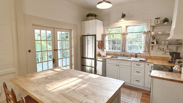 A kitchen with a wooden table, white cabinets, a stainless steel refrigerator, and a window with curtains. - NEW! Pine Cottage - Walk to Everything in Downtown Bryson City! (Bryson City)