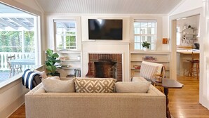 A cozy living room with a fireplace, a television mounted above it, and a dining area visible through an open door. - NEW! Pine Cottage - Walk to Everything in Downtown Bryson City! (Bryson City)