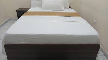 Una cama bien hecha con ropa blanca y almohadas, un cabecero de madera oscura y una mesita de noche con una lámpara y algunos objetos encima.