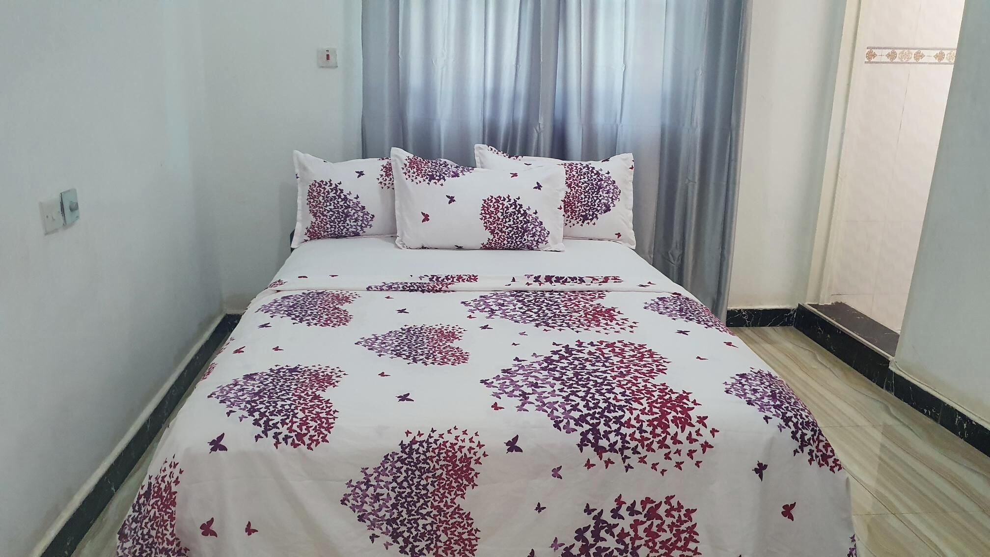 Een bed met een bloempatroon deken en passende kussens, een lichtgekleurde muur en een raam met grijze gordijnen.
