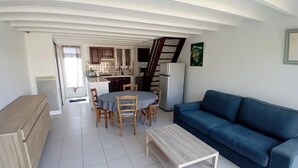 Een woonkamer met een blauwe bank, een houten salontafel en een eethoek met een ronde tafel en stoelen. Achtergrond toont een keuken met een koelkast en kasten.