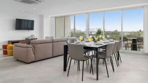 A modern living room with a dining area, large windows, and a flat-screen TV on the wall.