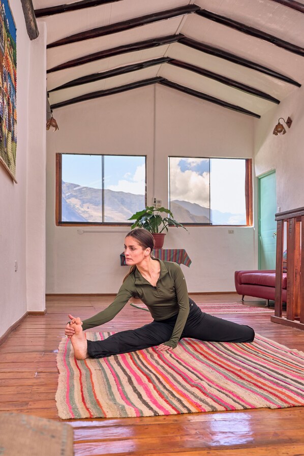 A person performing a yoga stretch on a striped rug in a room with wooden floors, a large window, and a plant.
