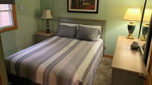 Una cama bien hecha con ropa de cama a rayas, una lámpara de noche, un cuadro enmarco con un paisaje, y una mesita con lámpara y florero.