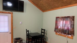 Una habitación con una mesa de comedor y sillas, un televisor montado en la pared, una ventana con cortinas y un techo de madera.
