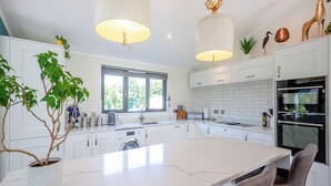 Une cuisine moderne avec des meubles blancs, un grand îlot central, des luminaires suspendus et une vue sur l’extérieur.