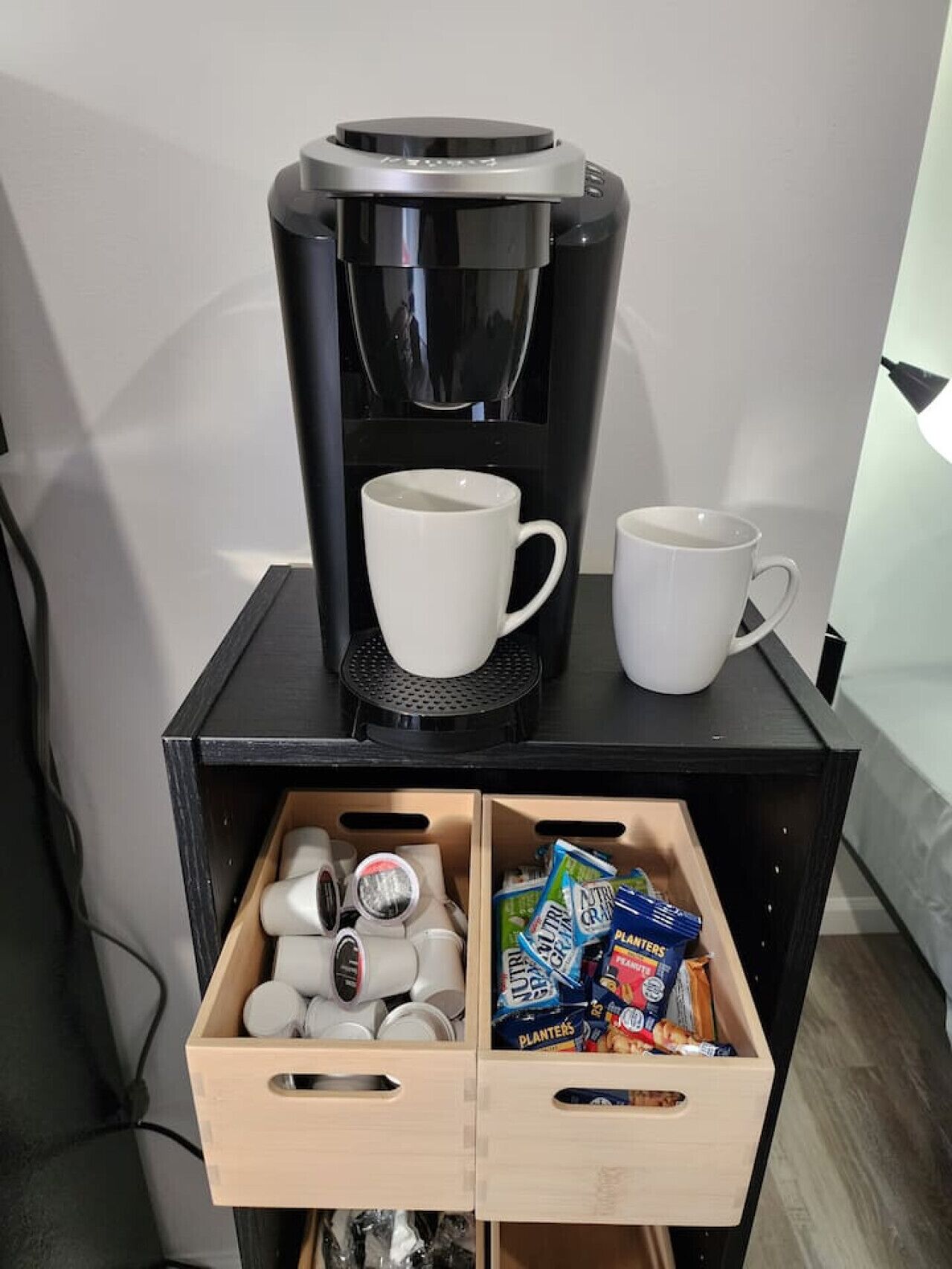 A coffee maker on a black stand with two white cups and a drawer below containing white cups and Planters brand snacks.