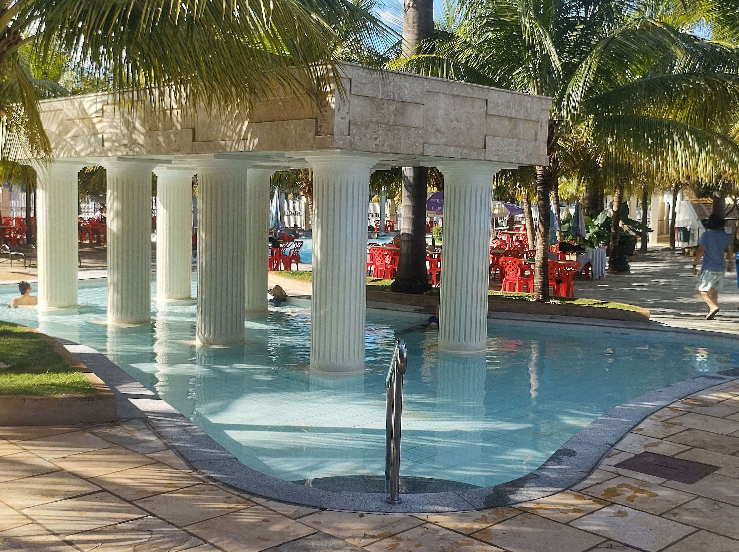 A pool area with a stone structure supported by columns, surrounded by palm trees and red chairs.