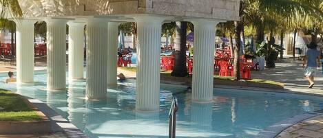 A pool area with a stone structure supported by columns, surrounded by palm trees and red chairs.