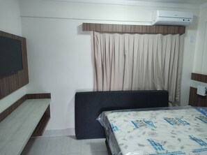 Una camera d'albergo con un letto, una TV a schermo piatto montata a parete, una finestra con tende e un condizionatore d'aria.