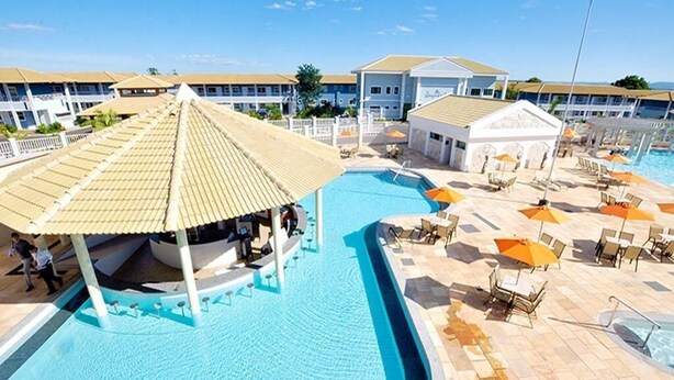Une piscine à bord incurvé, des chaises longues, des parasols et un bâtiment à la façade blanche en arrière-plan.