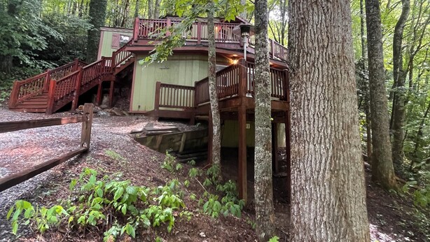 A wooden cabin with a balcony and stairs, surrounded by trees and a forested area.