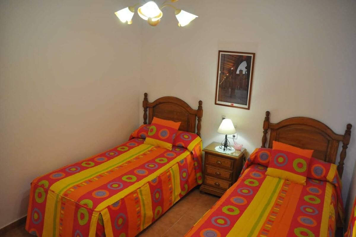 Ein Zimmer mit zwei Einzelbetten, beide mit hölzernen Kopfteilen und farbenfrohen Bettdecken. Zwischen den Betten steht ein Nachttisch mit einer Lampe, und an der Wand hängt ein gerahmtes Bild.