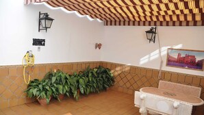 A corner of a room with a tiled floor, a white table with ornate legs, a potted plant, a yellow garden hose, and a framed picture on the wall. - Los galanes  (Villanueva de los Infantes)