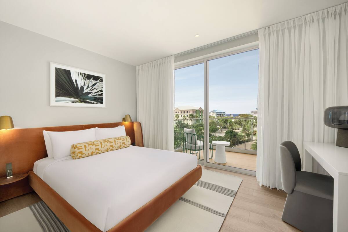 A hotel room with a large bed, a desk, a chair, a balcony with a view, and a painting on the wall.