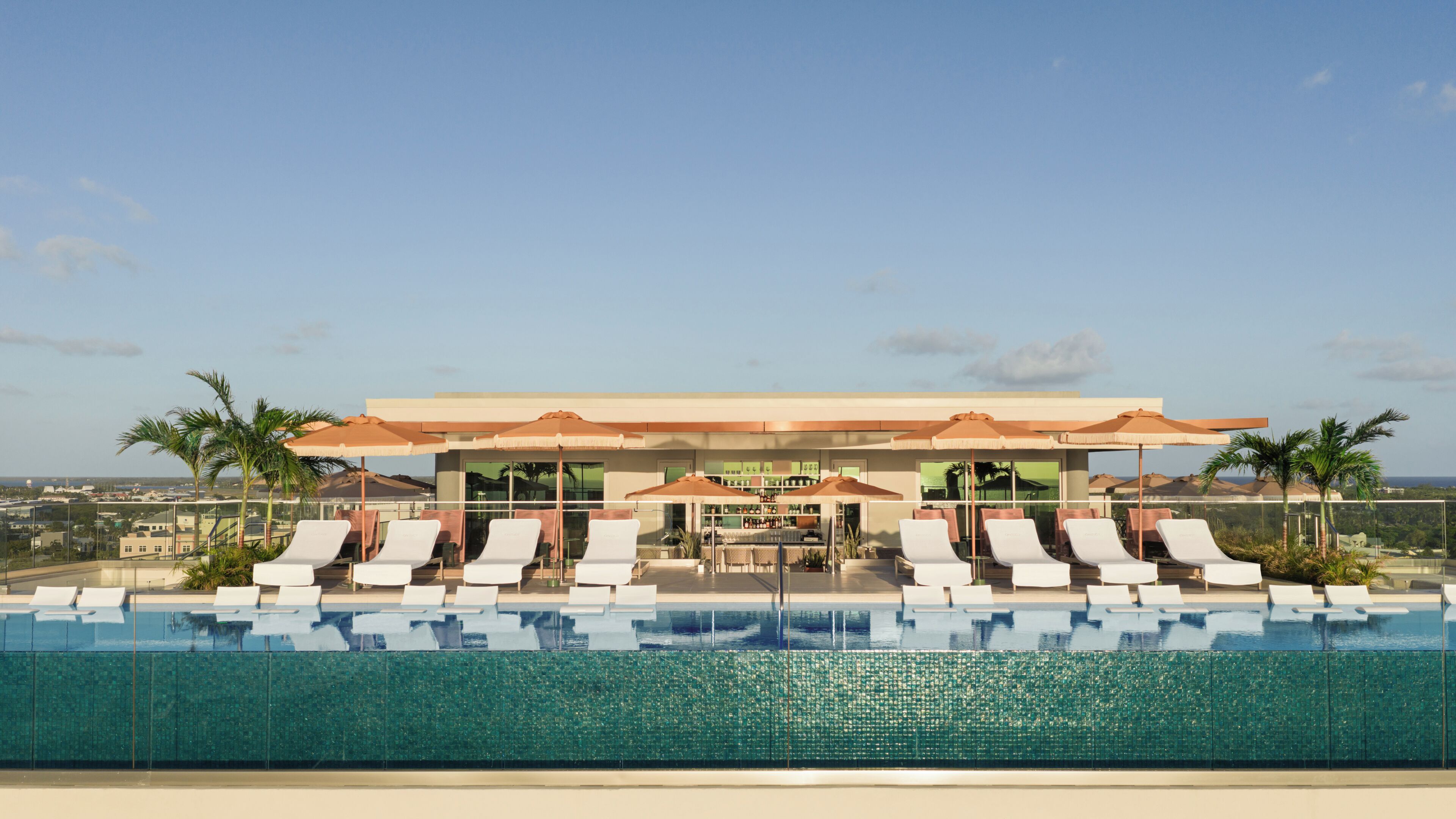 A rooftop infinity pool with lounge chairs and umbrellas, a bar area, and a view of the cityscape.