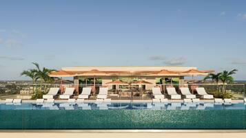 A rooftop infinity pool with lounge chairs and umbrellas, a bar area, and a view of the cityscape.