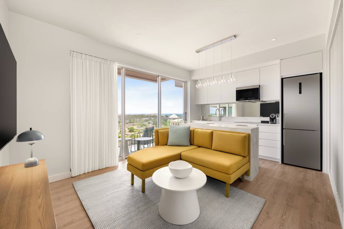 A modern kitchen with a built-in oven, a refrigerator, and a dining area with a yellow sofa and a round table.