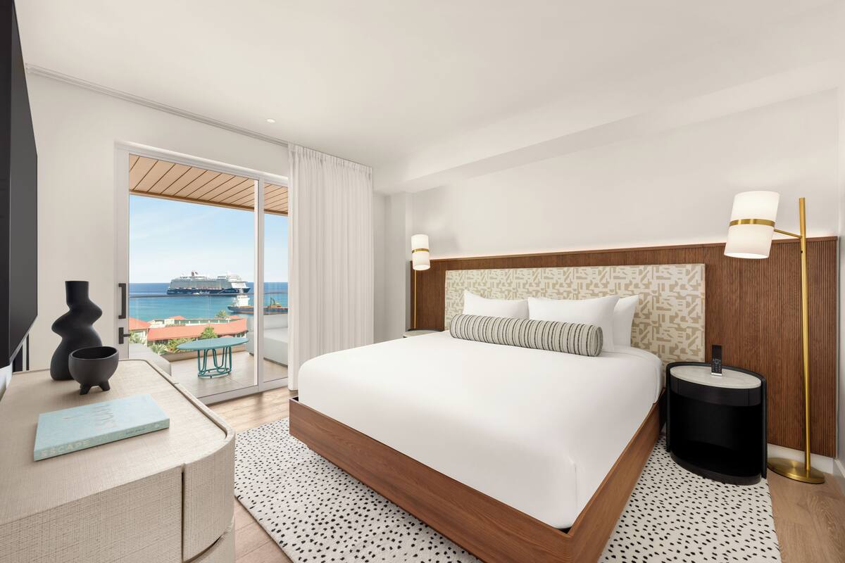 A modern hotel room with a large bed, a bedside table with a book, a nightstand, a lamp, and a view of the sea.
