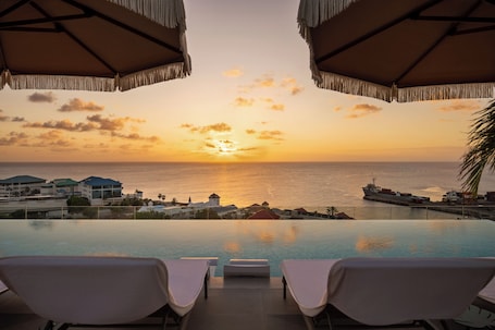 An infinity pool with lounge chairs and umbrellas, overlooking a sunset over the ocean with buildings in the background.