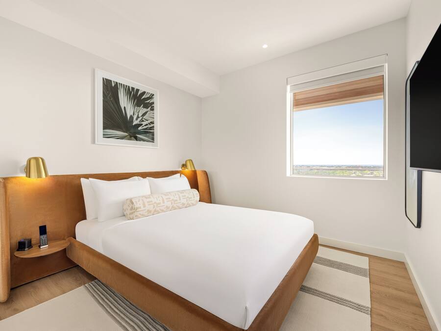 A modern hotel room with a large bed, bedside tables, a mounted TV, a window with a view, and a framed artwork on the wall.