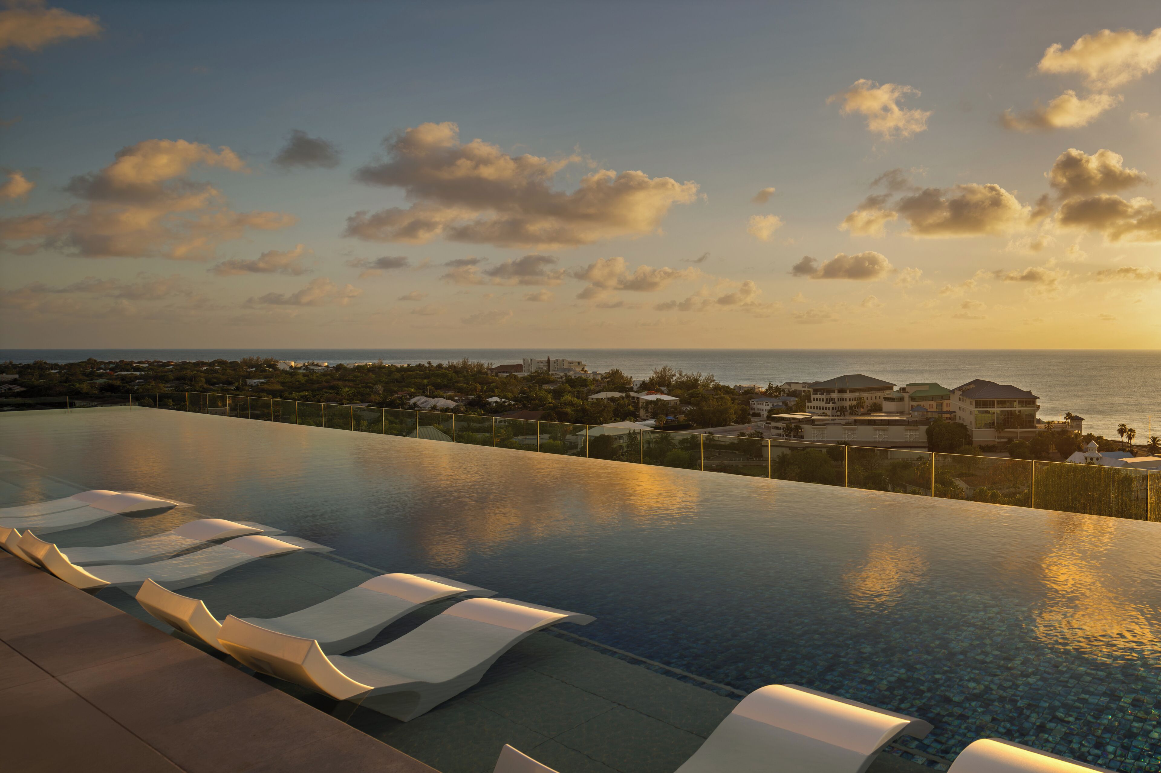 An infinity pool with lounge chairs, a view of the sea, and buildings in the background.