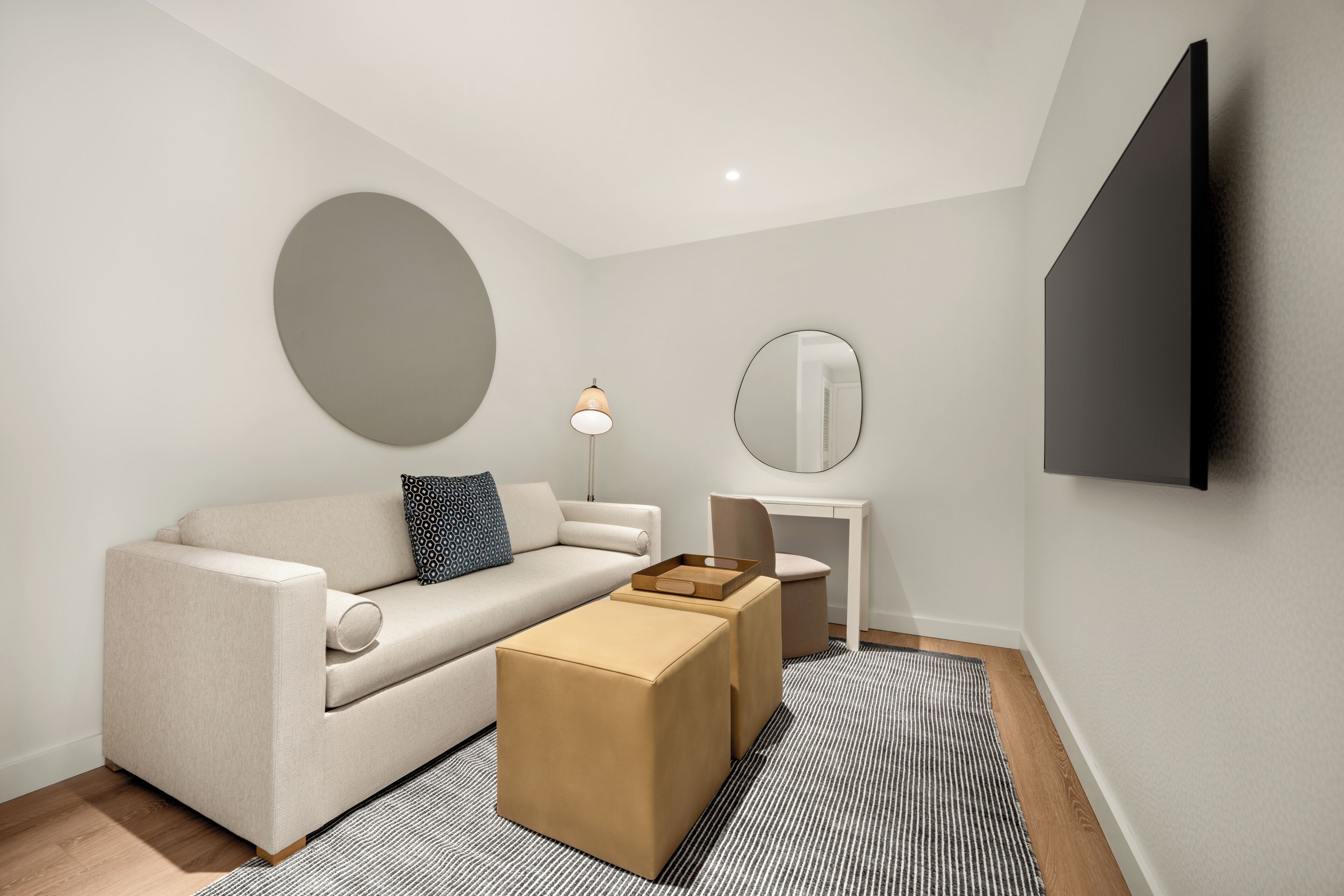 A modern living room with a sofa, ottoman, round mirror, and a flat-screen TV mounted on the wall.