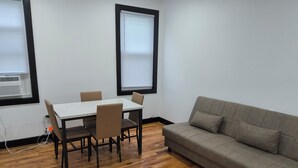 A room with a wooden floor, a white table with four chairs, a grey sofa, and two windows with blinds. - Spacious 4-bedroom, 4 beds 2 bath each, in lovely Newark with AC, WiFi. (Newark)