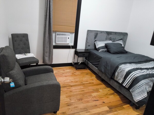 A bedroom with a bed, a chair, a nightstand, a window with blinds, and an air conditioning unit. - Spacious 4-bedroom, 4 beds 2 bath each, in lovely Newark with AC, WiFi. (Newark)