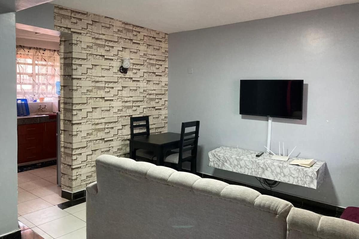 A living room with a sofa, a TV mounted on the wall, a marble side table, and a kitchen area visible through an open door.