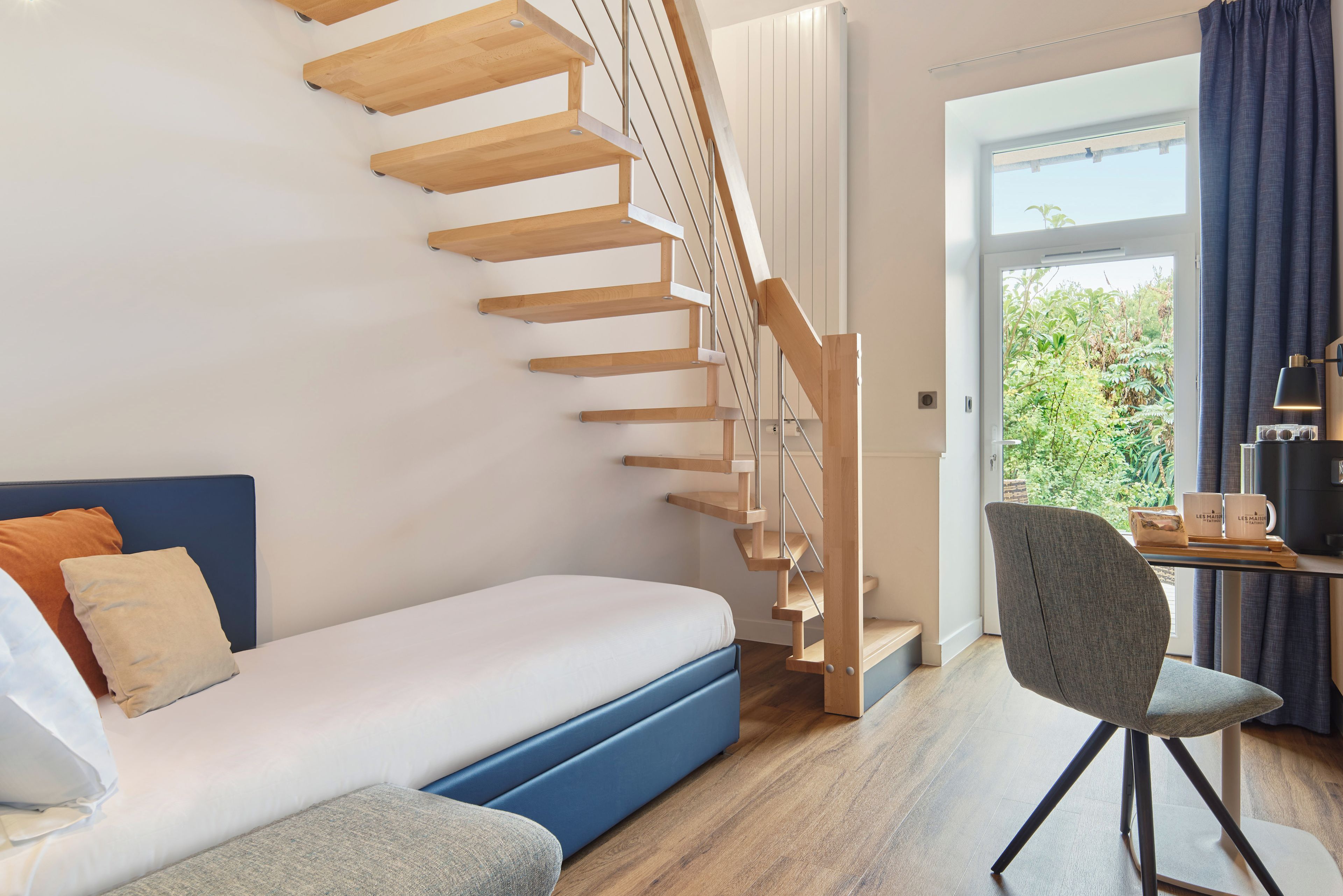 A modern room with a wooden staircase, a bed, a desk with a chair, and a view through a large window.