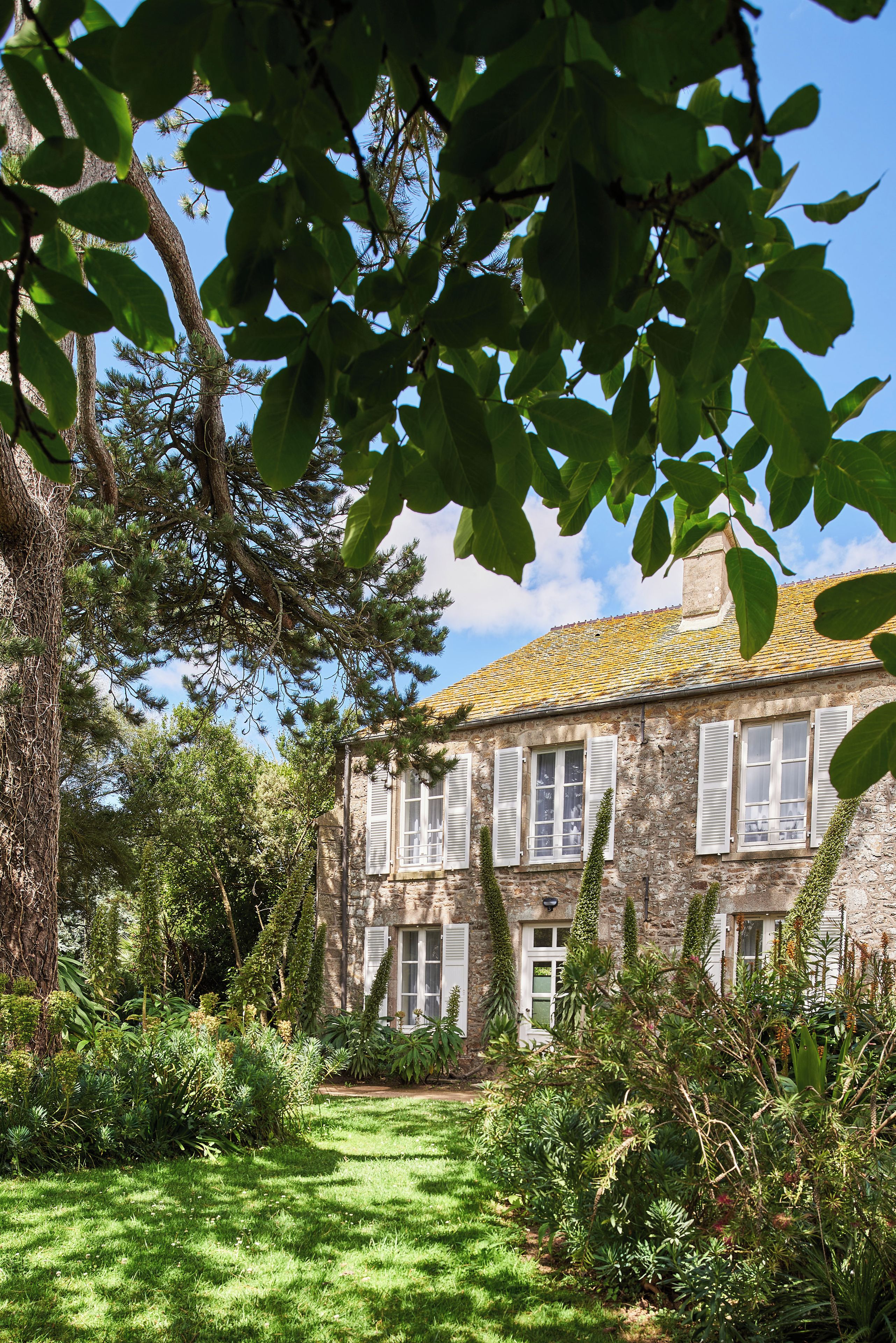 A stone building with multiple windows and shutters, surrounded by lush greenery and a well-maintained lawn.